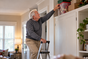 Elderly man standing on a step ladder to reach Christmas decoration boxes stored on a high shelf, highlighting potential fall hazards at home.